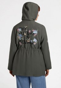 Grön huva jacka med blommönsterbroderi och text på baksidan. Elastisk midja, långa ärmar och en avslappnad passform. Avslappnad design.