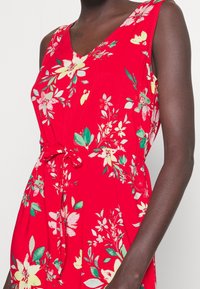 Bezramenné červené šaty s květinovým vzorem v žluté, růžové a zelené barvě, výstřih do V a opasek v pase, nošené osobou tmavé pleti, viditelný trup a ruka.