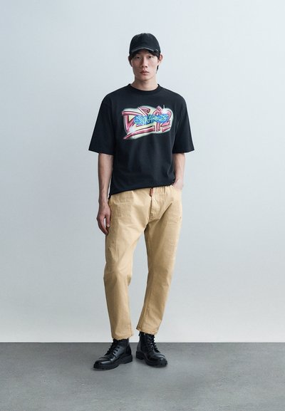 Jeune homme portant une casquette noire, un t-shirt graphique noir, un pantalon beige et des bottes à lacets noires, debout contre un mur clair uni.