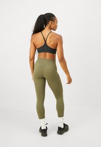 Kvinna med flätat hår i svart sportbh och olivgröna leggings står vänd bort på vit bakgrund.