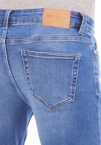 Jean en denim bleu avec une poche arrière, délavage léger, étiquette en cuir marron sur la ceinture avec l'inscription "ONLY & SON", détails de couture subtils.