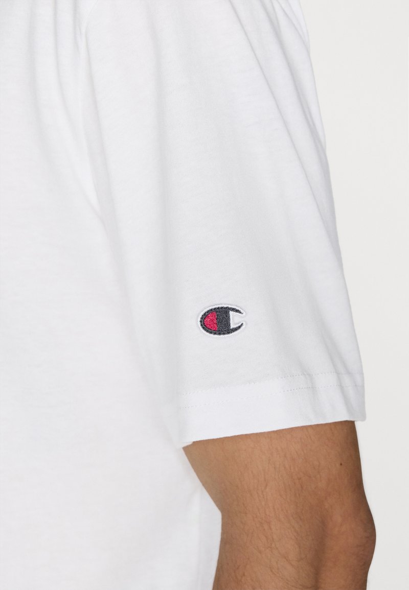 Camiseta blanca de manga corta hecha de algodón con un escote redondo. Presenta un pequeño logo bordado en rojo y negro en la manga izquierda.