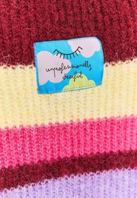 Kolorowa dzianina w paski w odcieniach czerwieni, różu, żółci i lawendy. Niebieska etykieta z chmurkami z napisem „nieprofesjonalnie wesoły”.