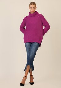 Fioletowy sweter dzianinowy z wysokim kołnierzem, luźnym krojem i prążkowaną fakturą. W połączeniu z niebieskimi dżinsami z frędzlami i ozdobnymi wykończeniami oraz czarnymi szpilkami.