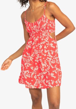 Femme portant une robe rouge à fleurs sans manches avec des bretelles fines et une découpe sur le côté, debout devant un fond blanc uni.