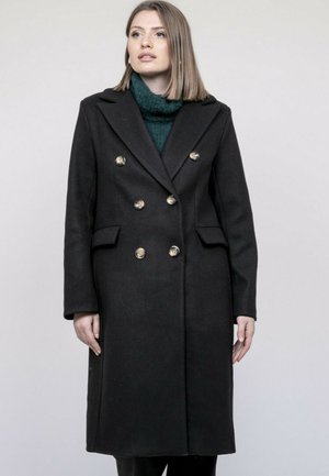 Femme portant un long manteau noir à double boutonnage avec des boutons dorés et un pull col roulé vert foncé, debout devant un fond clair.