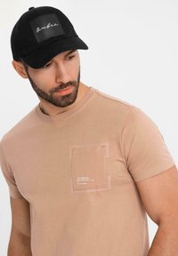 Béžové tričko s krátkým rukávem a přední kapsou, spojené s černou kšiltovkou s obdélníkovým výšivkou s bílým podpisem.