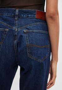 Tumši zilas džinsa bikses ar augstu jostasvietu, kontrastējošu oranžu šuvi, divām aizmugurējām kabatām un ādas uzliku uz jostasvietas.