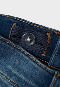 Denim pojas s tamnoplavom teksturiranom etiketom i transparentnim plastičnim dugmetom s cvjetnim uzorkom. Vidljiva kontrastna šav u žuto-smeđoj boji.