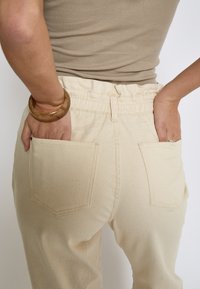 Pantaloni beige elasticizzati con due tasche posteriori, cintura elastica e cuciture visibili. Indossati con un braccialetto marrone al polso.