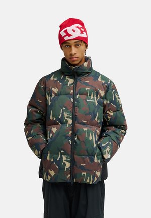 Jonge man draagt een groen-bruine camouflage donzen jas en een rode muts met wit logo, staand met handen in de jaszakken.