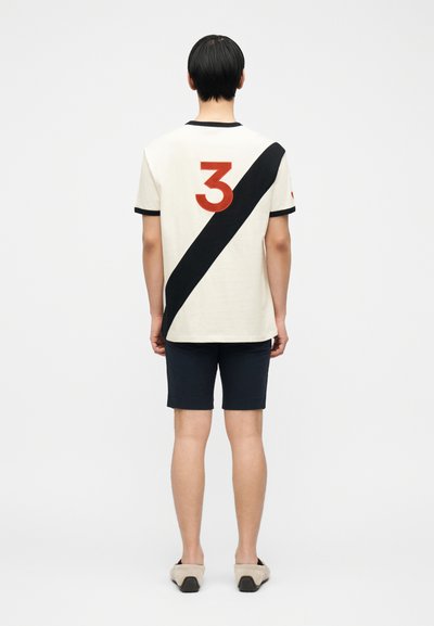 Άτομο με την πλάτη προς τα εμπρός, φοράει λευκό πουκάμισο με μαύρη διαγώνια γραμμή και κόκκινο αριθμό 3, σκούρο μπλε σορτς και μπεζ παπούτσια τύπου slip-on.