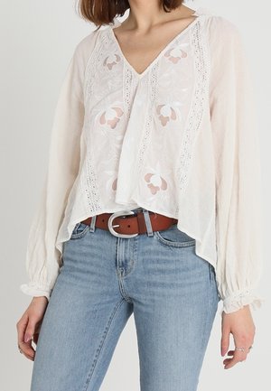 Kvinde iført en hvid broderet bluse med blonde detaljer, ballonsleeves, stoppet ned i lyseblå jeans med et brunt bælte.