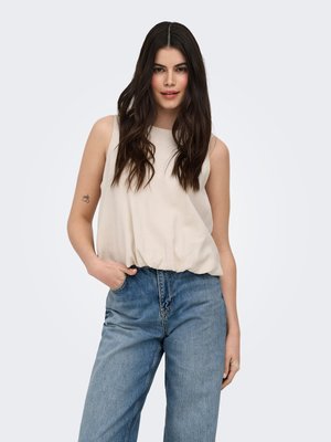 Femme aux longs cheveux foncés portant un top beige sans manches et un jean bleu ample, debout avec une main dans la poche sur un fond uni.