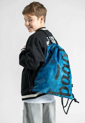 Zaino con cordino in tessuto tie-dye blu con scritte nere. Presenta cordini regolabili e una texture morbida. Indossato sopra un maglione nero.
