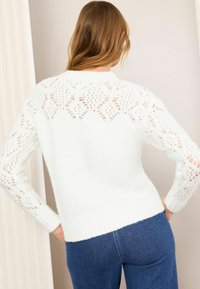 Weißer Strickpullover mit offenen Mustern an Ärmeln und Rücken, geripptem Saum und langen Ärmeln, kombiniert mit blauen Jeans.