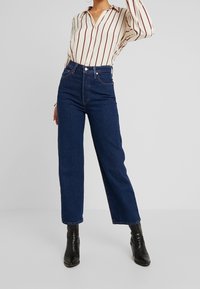 Vaqueros de cintura alta en azul con un corte recto, diseño clásico de 5 bolsillos y detalles de costura sutiles, combinados con una camisa de botones de rayas.