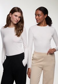 Due modelle indossano delle maglie bianche a maniche lunghe. Una modella ha dei pantaloni neri con tasche; l'altra indossa dei pantaloni della tuta beige con un elastico testurizzato in vita.