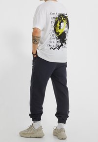 Osoba nosící bílé tričko s černým a žlutým grafickým motivem, černé teplákové kalhoty, bílé ponožky a béžové tenisky, stojící s rukama v kapsách.