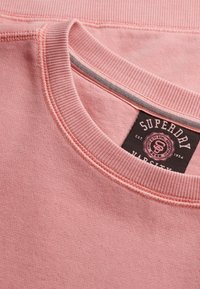 Różowa bluza z ribbowym dekoltem, gładką teksturą oraz czarną etykietą Superdry z napisem "Varsity". Subtelne detale w szyciu.