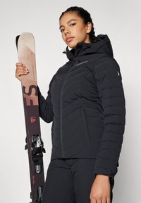 Black puffer jacket with zip closure and hood, featuring horizontal stitching. Person holds skis with colorful detailing and bindings.