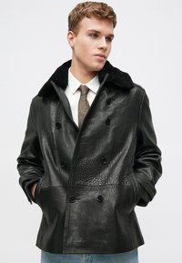 Veste en cuir noir à double boutonnage avec une finition texturée, dotée d'un col en fourrure noire et de poches latérales, portée sur une chemise blanche et une cravate.
