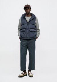 Chaleco de plumón navy con cuello alto y botones de presión, sudadera gris debajo, zapatos verdes y marrones, pantalones sueltos teal.