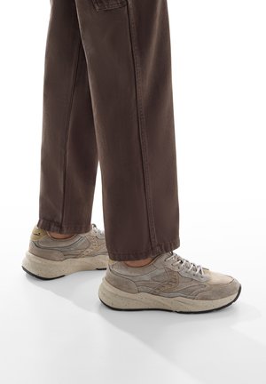 Sneaker beige con tomaia texturizzata, materiali scamosciati e in mesh, con suole in gomma maculate e accenti grigi, abbinate a pantaloni marroni.