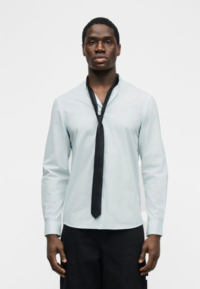 Νέος άνδρας με σκούρο δέρμα που φοράει ανοιχτό μπλε πουκάμισο μακρυμάνικο και χαλαρά δεμένη μαύρη γραβάτα, στέκεται μπροστά σε απλό λευκό φόντο.