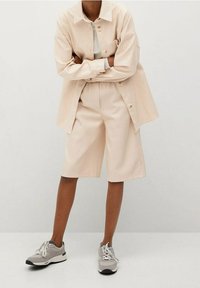 Osoba ubrana w beżową koszulę z długim rękawem oraz dopasowane szorty do kolan w tym samym kolorze, w jasnoszarych tenisówkach, stojąca z założonymi rękami na tle białym.