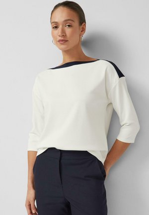 Kobieta w białej bluzce z granatowymi wstawkami na ramionach i granatowych spodniach, stojąca na jasnoszarym tle z rękami w kieszeniach.