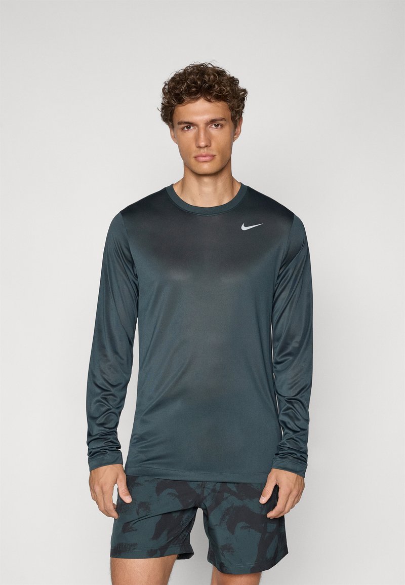 Langærmet atletisk top i mørkegrønt stof med en glat tekstur, der har en subtil gradient og et lille hvidt Nike-logo på brystet.