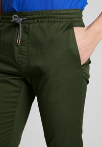 Pantalon vert léger avec une texture lisse, taille élastique, cordon de serrage gris, et poches latérales. Convient pour une tenue décontractée.