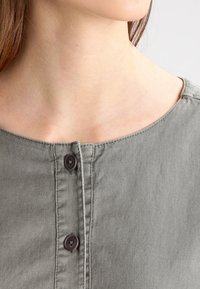 Harmaa, pehmeä kangas yläosa, jossa on pyöreä kaula-aukko, kaksi tummanruskeaa nappia ja huomaamaton ompelu napituksessa.