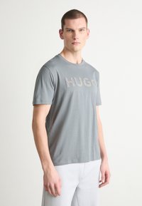 Pelēka kokvilnas T-krekls ar īsām piedurknēm un apaļu kakla izgriezumu, kurā uz krūtīm ir vārds "HUGO" kontrastējošā, tekstūrētā dizainā.