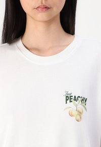 Valge puuvillane T-särk, millel on trükitud disain, millel on tekst "Just Peachy" ning graafika virsikutest roheliste lehtedega. Ümar kaelus.