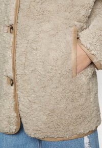 Manteau en peau lainée beige clair avec une surface douce et texturée et une garniture beige, présentant des boutons visibles et une poche latérale. Coupe décontractée conçue pour la chaleur.