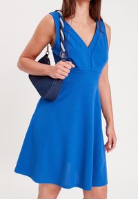 Robe bleue sans manches avec un décolleté en V et un corsage ajusté, jupe fluide et texture lisse, assortie à un petit sac à main sombre.