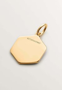 Charm hexagonal de oro con una superficie lisa y reflectante. Grabado con la palabra "aristocrazy". Incluye un pequeño lazo circular de sujeción.