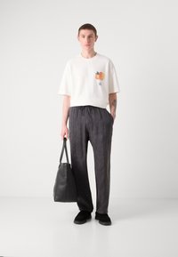 Hvit kortermet t-skjorte med fruktgrafikk, kombinert med løstsittende mørkegrå bukser; modellen holder en svart skinn tote bag.