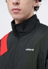 Μαύρο αθλητικό μπλουζάκι με ψηλό γιακά, κόκκινη λεπτομέρεια και λευκό λογότυπο adidas. Ορατό υφασμάτινο σχέδι�ο και κλείσιμο με ολόκληρο φερμουάρ.