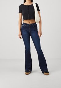 Schwarzes, kurzes T-Shirt mit Knopfdetails, dunkelblaue Schlagjeans und schwarze Schuhe an einer Person, die eine weiße Umhängetasche hält.