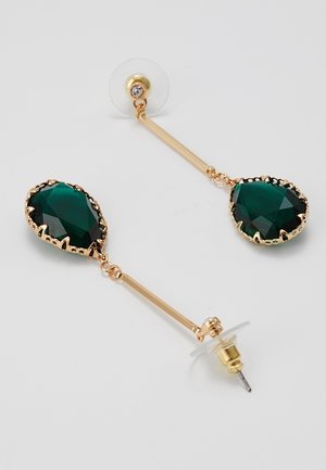 Kultaiset pisarakorvakorut, joissa pitkät varret ja suuret tummanvihreät, monisärmäiset pisaranmuotoiset jalokivet kynsiasetuksissa valkoisella taustalla.