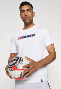 Hvid kortærmet T-shirt med et blåt og rødt "Nike Paris SG" logo. Holdes i hånden er en hvid fodbold med sorte og røde accenter samt et struktureret mønster.