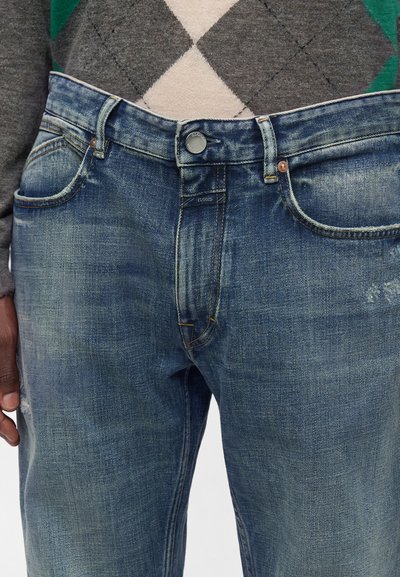 Jean en denim bleu légèrement usé avec braguette à boutons et fermeture éclair, associé à un pull gris à motif losanges.