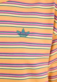 Randig gul t-shirt med rosa, grön och vita linjer; har en broderad teal-logga nära halsöppningen. Mjuk bomullstyg.