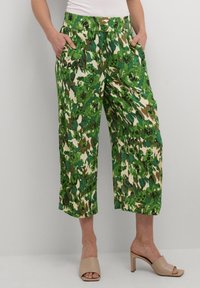 Pantaloni cropped a stampa floreale verde con una cintura elastica, due tasche laterali e una texture fluida, abbinati a mules beige con punte aperte.