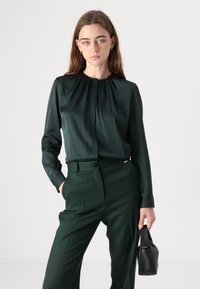 Young woman in dark green blouse and pants holding a small black handbag, standing against a plain light background.