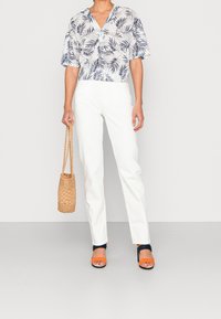 Blouse blanche courte avec motif de feuilles bleues, associée à un pantalon blanc à jambes droites. Accessoirisée avec un sac tressé couleur tan et des talons orange.
