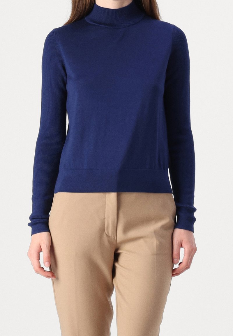 Femme portant un pull à col roulé bleu marine et un pantalon beige, debout devant un fond uni.